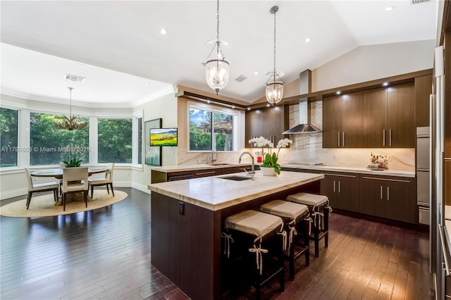 kitchen with a chandelier, dark wood finished floors, stovetop, a kitchen island with sink, and wall chimney range hood