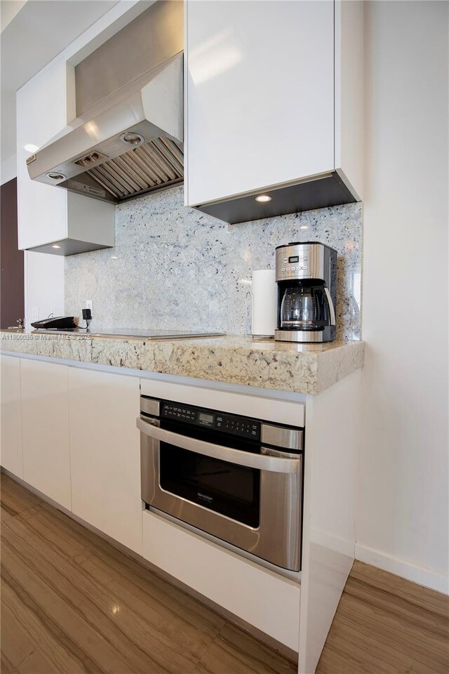 9705 Collins Ave Unit 1805N, Bal Harbour, FL 33154 - kitchen with wall chimney exhaust hood, stainless steel oven, white cabinetry, wood finished floors, and tasteful backsplash