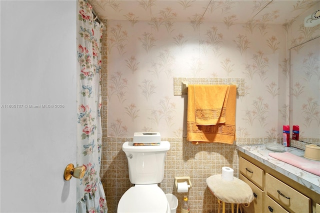 bathroom with toilet, wallpapered walls, wainscoting, vanity, and tile walls