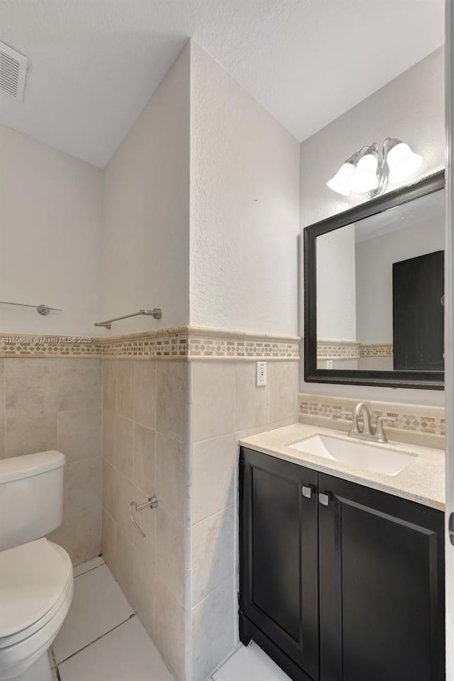 bathroom with tile walls, toilet, tile patterned floors, vanity, and a wainscoted wall
