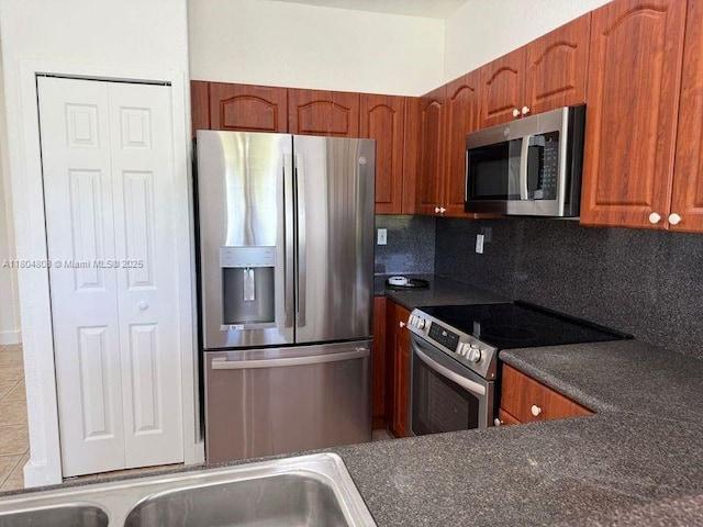 kitchen featuring stainless steel appliances, dark countertops, decorative backsplash, brown cabinetry, and a sink