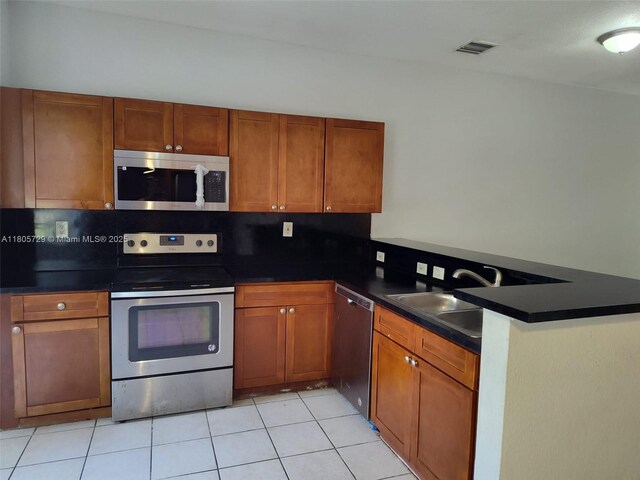 kitchen with appliances with stainless steel finishes, a peninsula, brown cabinetry, dark countertops, and a sink