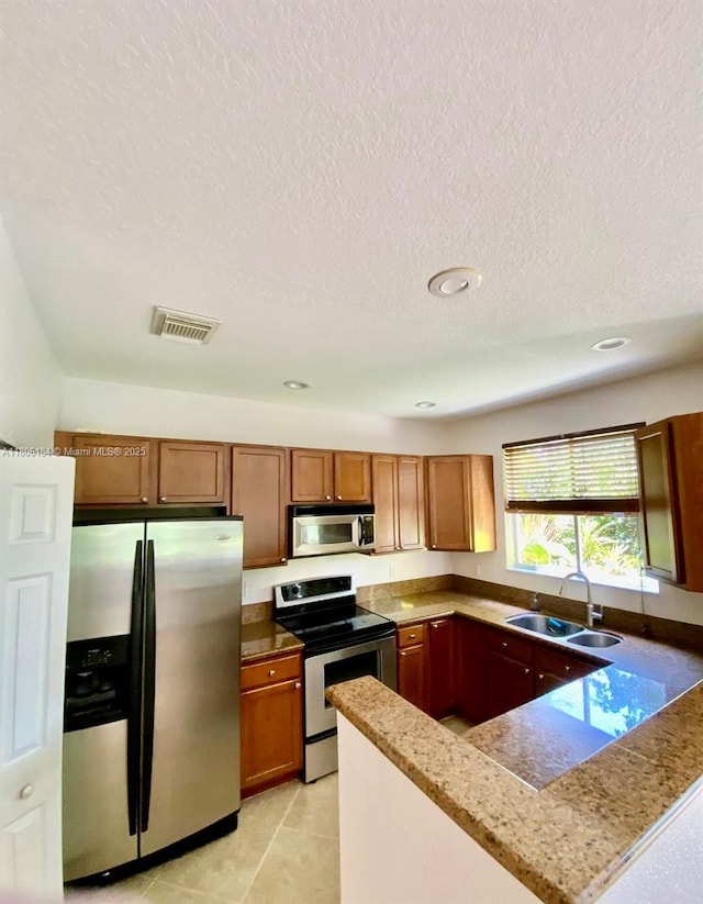 kitchen with appliances with stainless steel finishes, a sink, brown cabinetry, a textured ceiling, and a peninsula