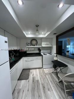801 Three Islands Blvd Unit 112, Hallandale Beach, FL 33009 - kitchen with white appliances, a raised ceiling, white cabinets, light wood-style floors, and dark countertops