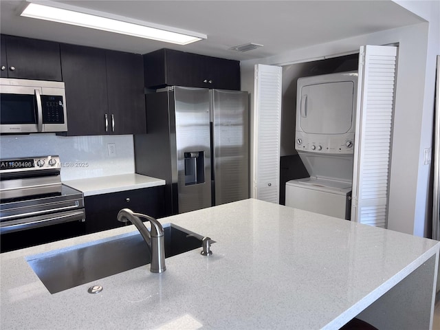 kitchen with appliances with stainless steel finishes, dark cabinets, stacked washer / drying machine, a sink, and light stone countertops
