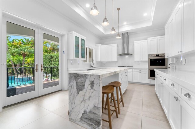1210 95th St, Bay Harbor Islands, FL 33154 - kitchen with wall chimney range hood, a tray ceiling, stainless steel double oven, light tile patterned flooring, and ornamental molding