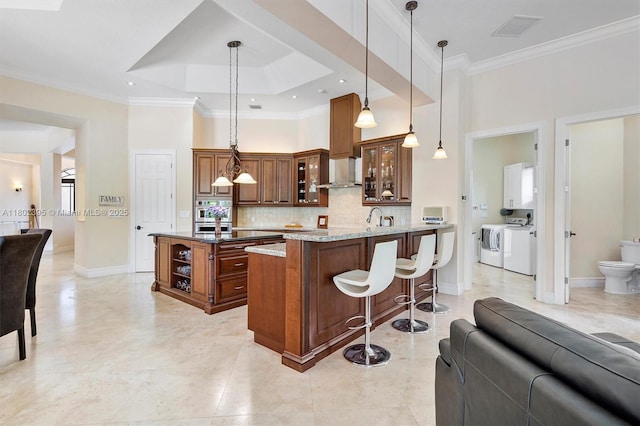 20120 SW 133rd Ave, Miami, FL 33177 - kitchen featuring washer and dryer, wall chimney exhaust hood, brown cabinets, a kitchen breakfast bar, and backsplash