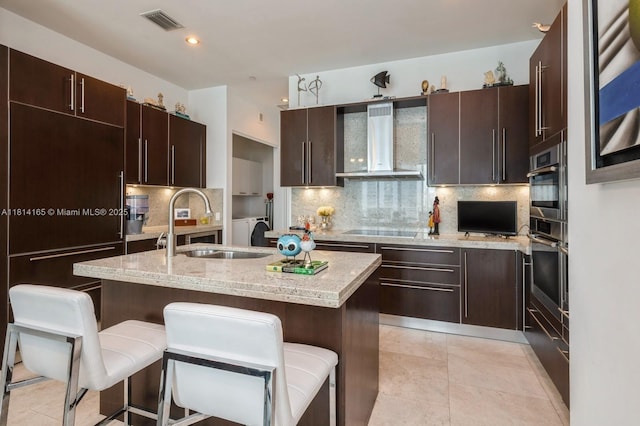 900 Brickell Key Blvd Unit 1702, Miami, FL 33131 - kitchen with wall chimney range hood, black electric stovetop, a breakfast bar area, a center island with sink, and decorative backsplash