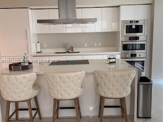 300 Sunny Isles Blvd Unit 4-1407, Sunny Isles Beach, FL 33160 - kitchen featuring wall chimney range hood, a breakfast bar, white cabinetry, double oven, and light countertops