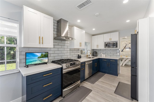 kitchen with appliances with stainless steel finishes, wall chimney range hood, blue cabinetry, white cabinets, and decorative backsplash