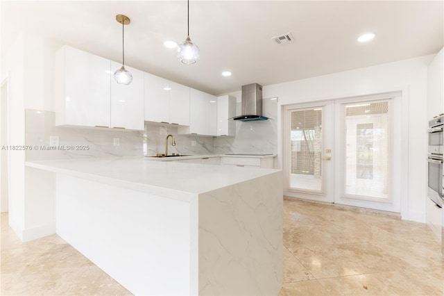 kitchen with wall chimney exhaust hood, white cabinets, a peninsula, decorative backsplash, and hanging light fixtures