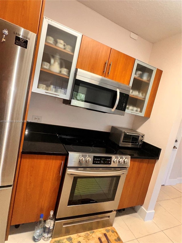 18800 NE 29th Ave Unit 412, Aventura, FL 33180 - kitchen featuring appliances with stainless steel finishes, brown cabinetry, light tile patterned floors, glass insert cabinets, and dark countertops