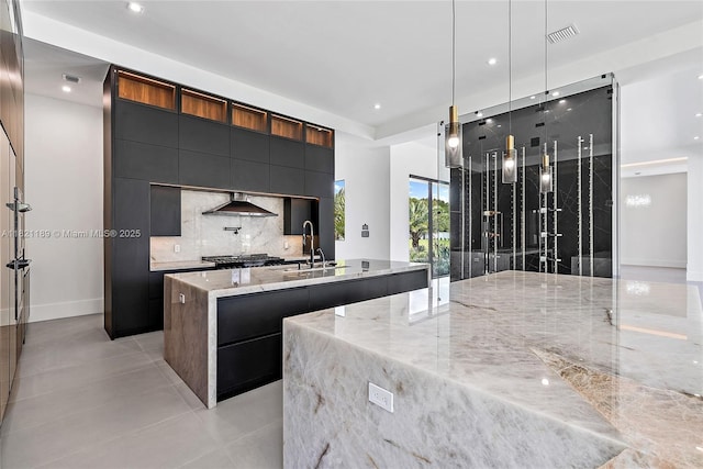 9741 SE Gomez Ave, Hobe Sound, FL 33455 - kitchen with modern cabinets, a center island with sink, tasteful backsplash, light stone countertops, and wall chimney range hood