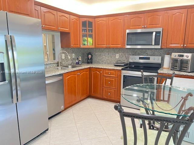 905 NW 107th Ave, Pembroke Pines, FL 33026 - kitchen with appliances with stainless steel finishes, backsplash, light tile patterned floors, brown cabinetry, and light stone countertops