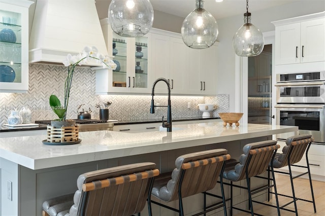 9369 Crestview Cir, Palm Beach Gardens, FL 33412 - kitchen with double oven, backsplash, white cabinets, glass insert cabinets, and hanging light fixtures