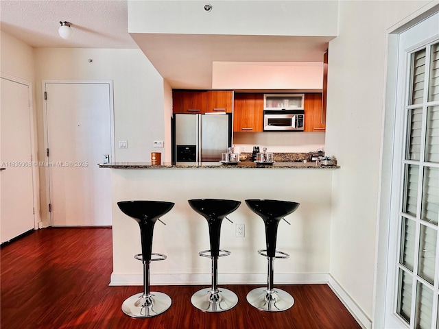 9066 SW 73rd Ct Unit 1409, Miami, FL 33156 - kitchen with stainless steel appliances, brown cabinetry, dark wood-style flooring, a breakfast bar area, and dark stone counters