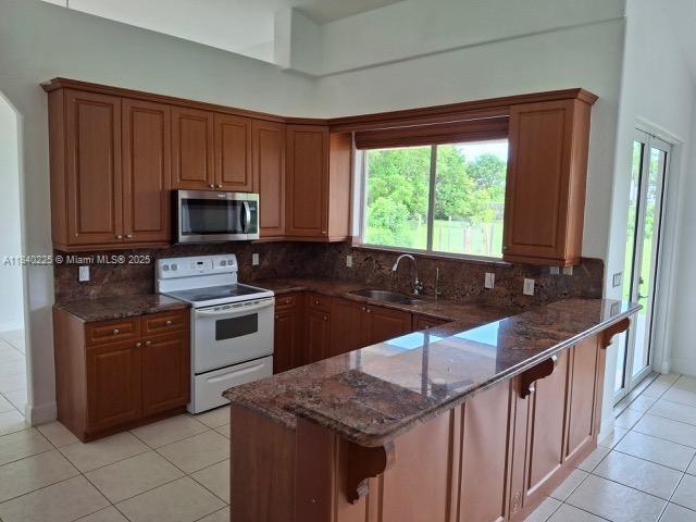 16961 SW 213th Ln, Miami, FL 33187 - kitchen with stainless steel microwave, white electric range, a peninsula, light tile patterned floors, and dark stone countertops