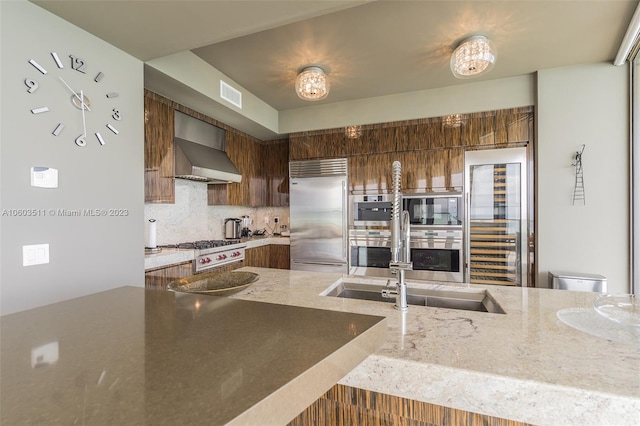 kitchen featuring wall chimney range hood, stainless steel appliances, sink, light stone counters, and backsplash