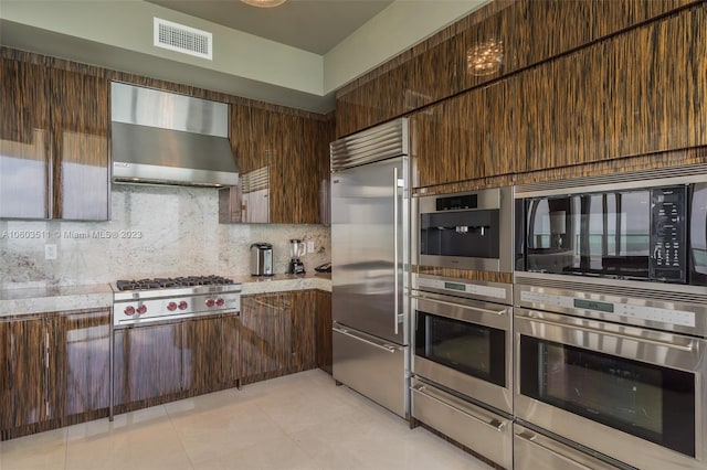 kitchen featuring dark brown cabinets, tasteful backsplash, wall chimney range hood, light tile patterned floors, and built in appliances