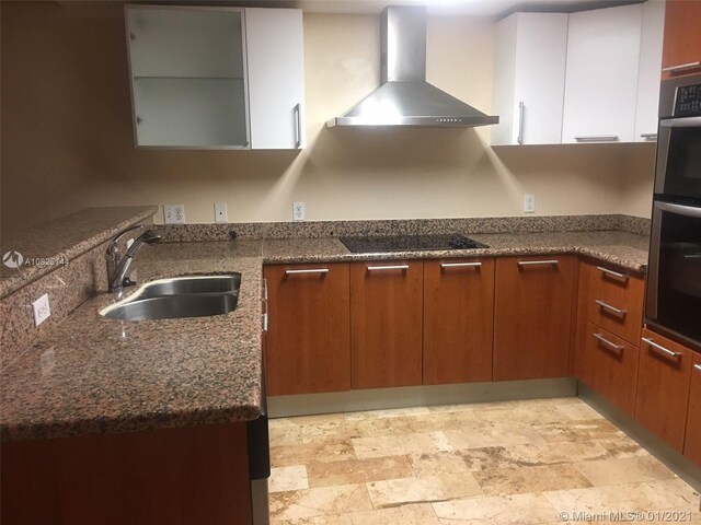 kitchen featuring dark stone countertops, wall chimney exhaust hood, a sink, brown cabinets, and black electric stovetop