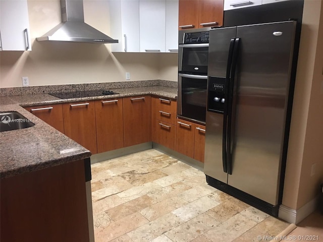 kitchen featuring dark stone countertops, stone finish floor, wall chimney exhaust hood, brown cabinetry, and appliances with stainless steel finishes