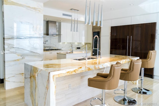 kitchen featuring light wood-type flooring, a kitchen island with sink, sink, hanging light fixtures, and wall chimney exhaust hood