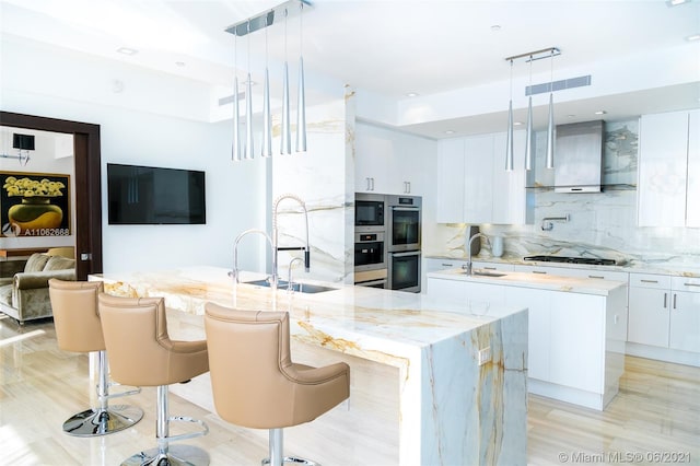 kitchen with hanging light fixtures, a center island with sink, wall chimney range hood, and white cabinets