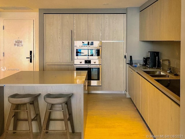 kitchen featuring light brown cabinetry, sink, stainless steel double oven, a breakfast bar, and light wood-type flooring