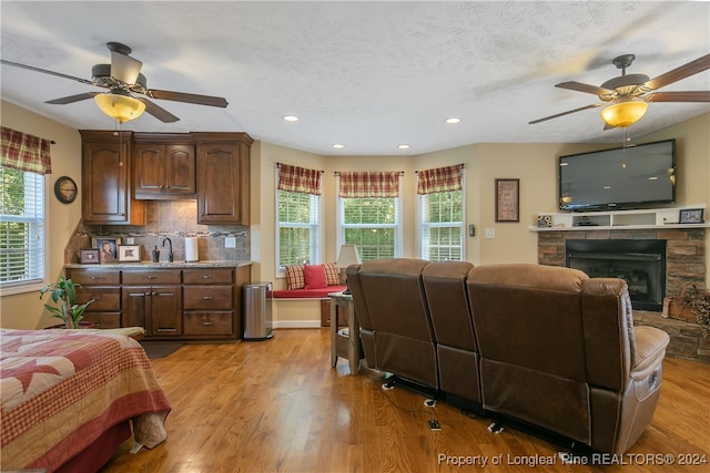 bedroom with a textured ceiling, light wood-type flooring, a stone fireplace, and ceiling fan