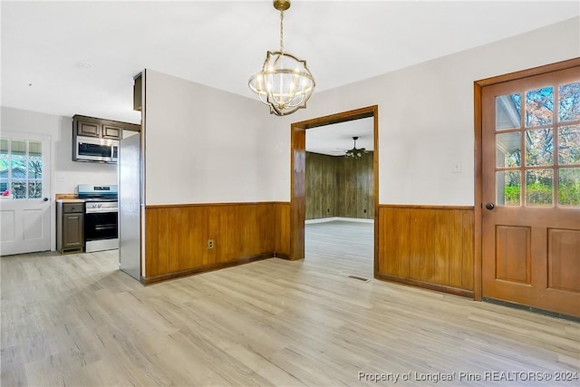 unfurnished dining area with wood walls, ceiling fan with notable chandelier, and light hardwood / wood-style floors