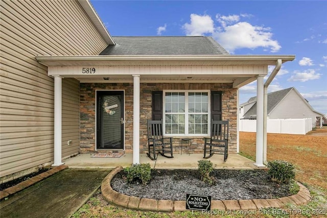 property entrance with stone siding, a porch, a shingled roof, and fence