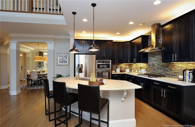 kitchen featuring hanging light fixtures, wall chimney range hood, stainless steel appliances, decorative columns, and a center island with sink