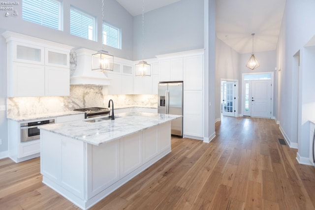 kitchen with a center island with sink, white cabinets, glass insert cabinets, appliances with stainless steel finishes, and custom exhaust hood