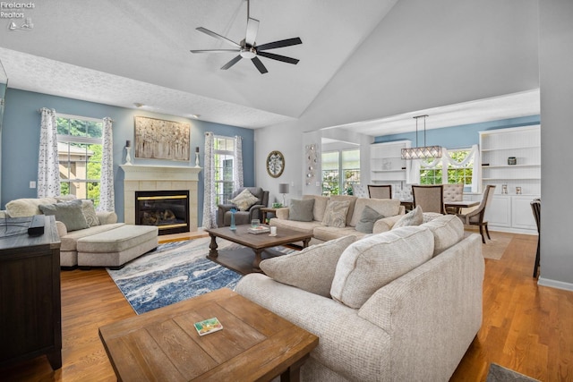 living room with light hardwood / wood-style flooring, ceiling fan with notable chandelier, a tile fireplace, high vaulted ceiling, and plenty of natural light