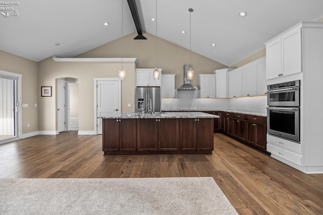 kitchen featuring an island with sink, stainless steel appliances, arched walkways, wall chimney range hood, and decorative backsplash