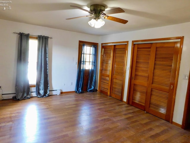 unfurnished bedroom featuring a baseboard heating unit, ceiling fan, wood-type flooring, and multiple closets