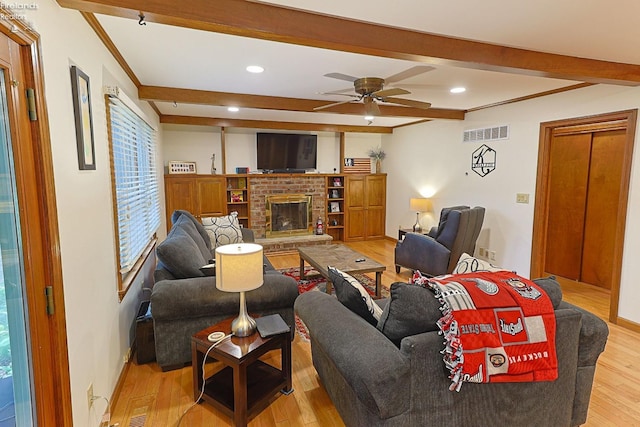 living room with light wood-type flooring, beamed ceiling, a fireplace, crown molding, and ceiling fan