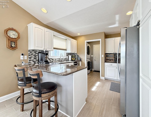 kitchen featuring stainless steel refrigerator, dark stone counters, stacked washer and clothes dryer, kitchen peninsula, and white cabinets