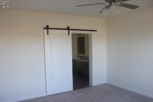 unfurnished bedroom with a barn door, dark colored carpet, ensuite bathroom, and ceiling fan