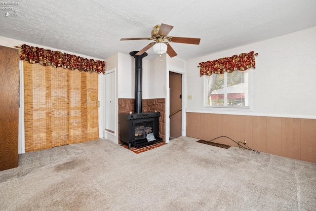 unfurnished living room with a wood stove, a textured ceiling, ceiling fan, wooden walls, and carpet