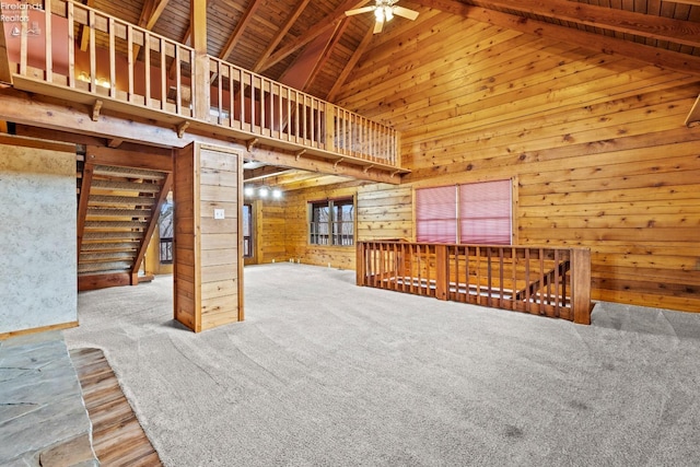 unfurnished living room with carpet flooring, ceiling fan, wooden walls, high vaulted ceiling, and beamed ceiling