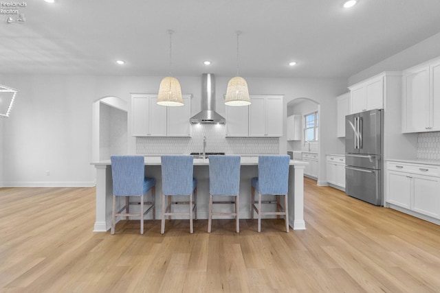 kitchen featuring hanging light fixtures, wall chimney range hood, stainless steel fridge, an island with sink, and white cabinets