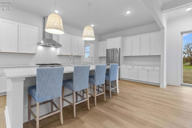 kitchen with hanging light fixtures, wall chimney range hood, a kitchen island with sink, a breakfast bar, and white cabinets