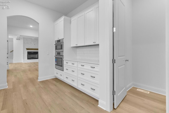 kitchen featuring white cabinetry, multiple ovens, decorative backsplash, a fireplace, and light wood-type flooring