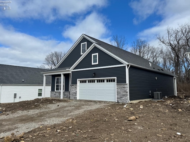 view of front of home featuring roof with shingles, dirt driveway, an attached garage, central AC, and stone siding