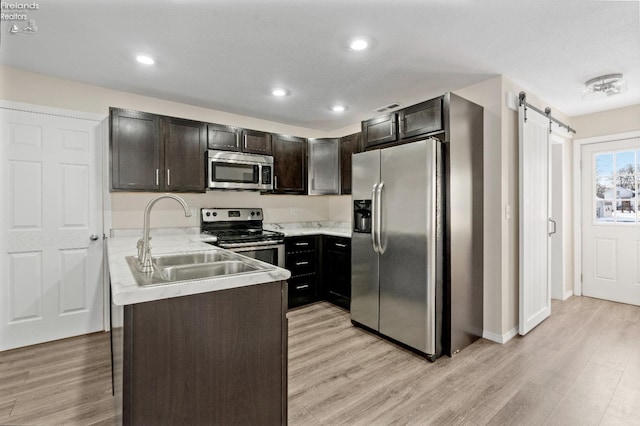 kitchen with appliances with stainless steel finishes, dark brown cabinetry, sink, a barn door, and light hardwood / wood-style floors