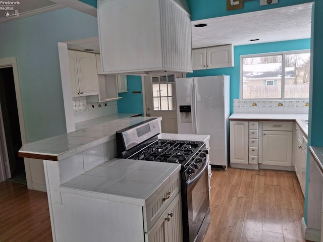 kitchen with kitchen peninsula, white refrigerator with ice dispenser, stainless steel range with gas cooktop, tile countertops, and light hardwood / wood-style floors