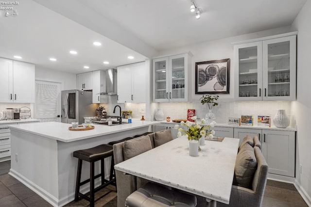 kitchen featuring wall chimney range hood, stainless steel fridge, a breakfast bar, a kitchen island with sink, and white cabinetry