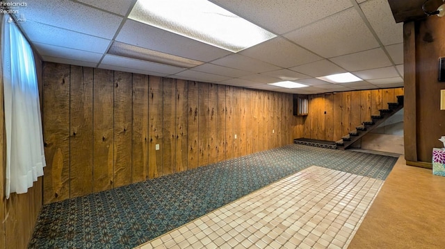 basement with a drop ceiling, light carpet, and wood walls