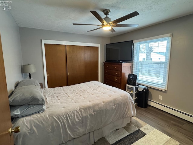 bedroom featuring dark wood-type flooring, a textured ceiling, a closet, ceiling fan, and a baseboard heating unit